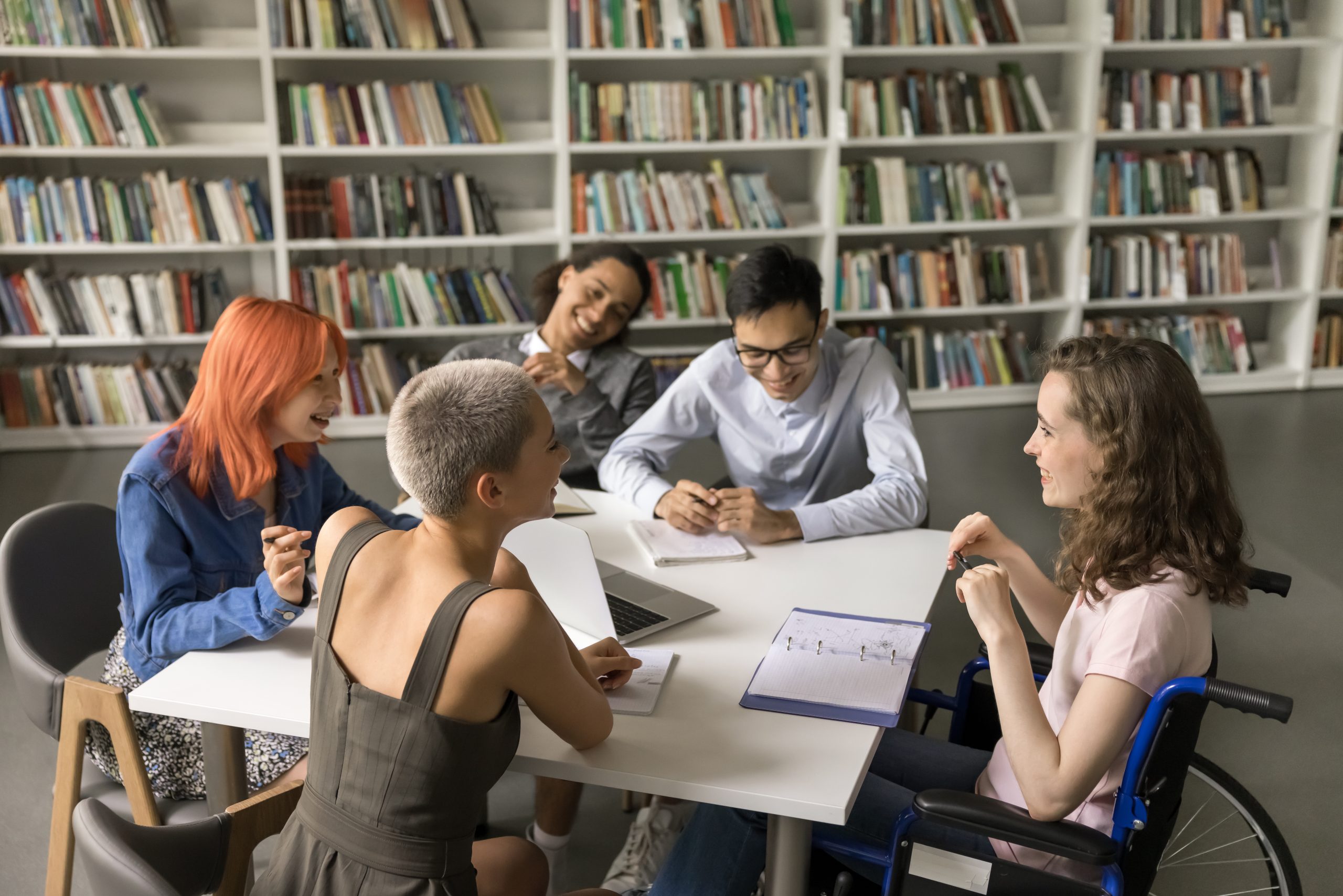 Group of students studying in library|This image shows people looking ahead and smiling|5 people sitting around a table smiling. In the background there are several shelves of books.