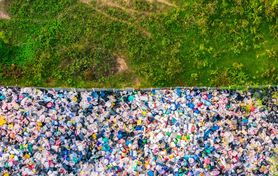 A striking top-down aerial view showing the sharp contrast between vibrant green grass and a chaotic pile of plastic waste, visually illustrating the environmental impact of consumerism on untouched nature.