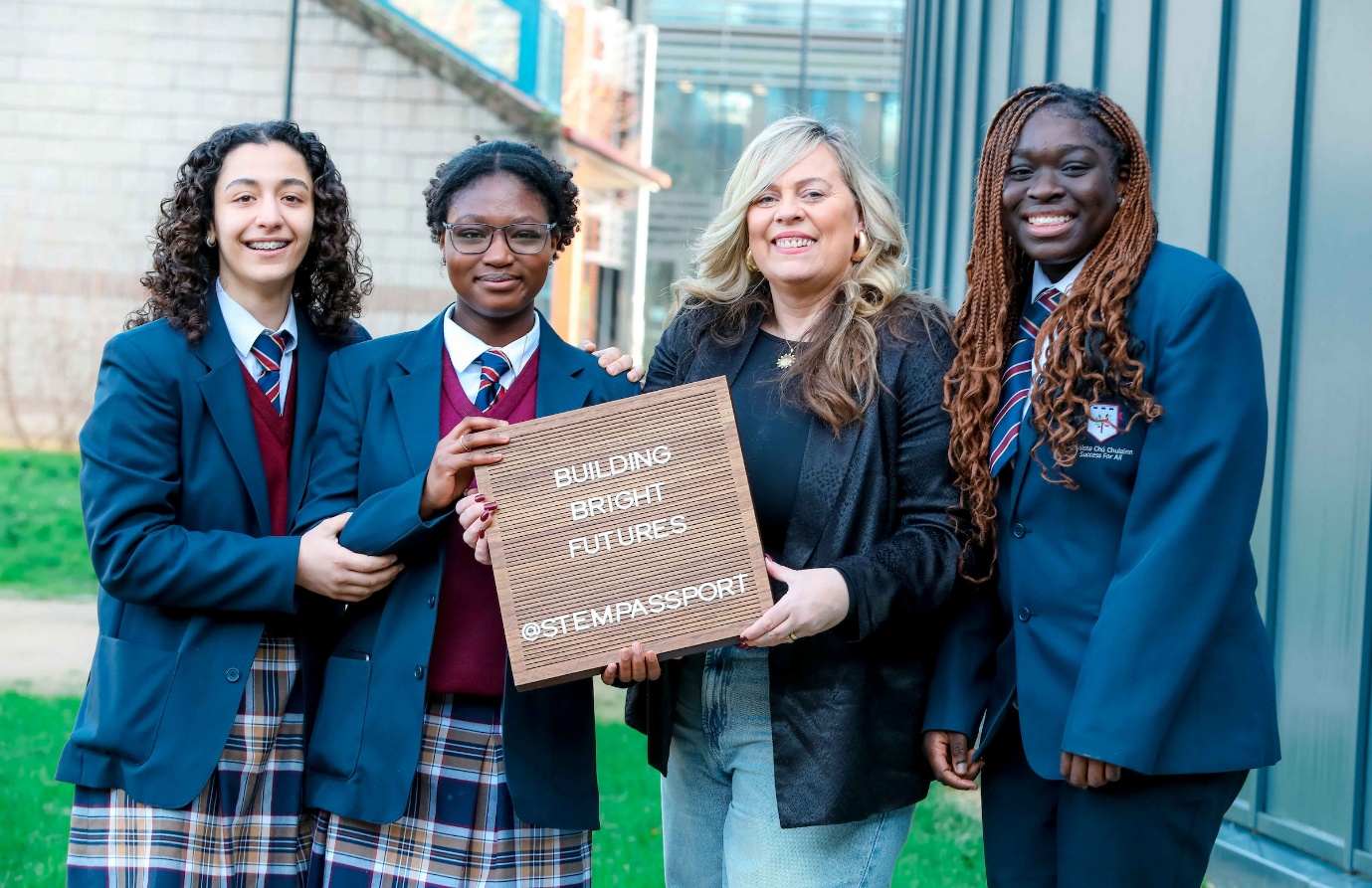 Four people stand together outdoors beside a modern building, with three wearing school uniforms and one wearing a coat. They are holding a wooden sign that reads “Building Brighter Futures #STEMPASSPORT.”