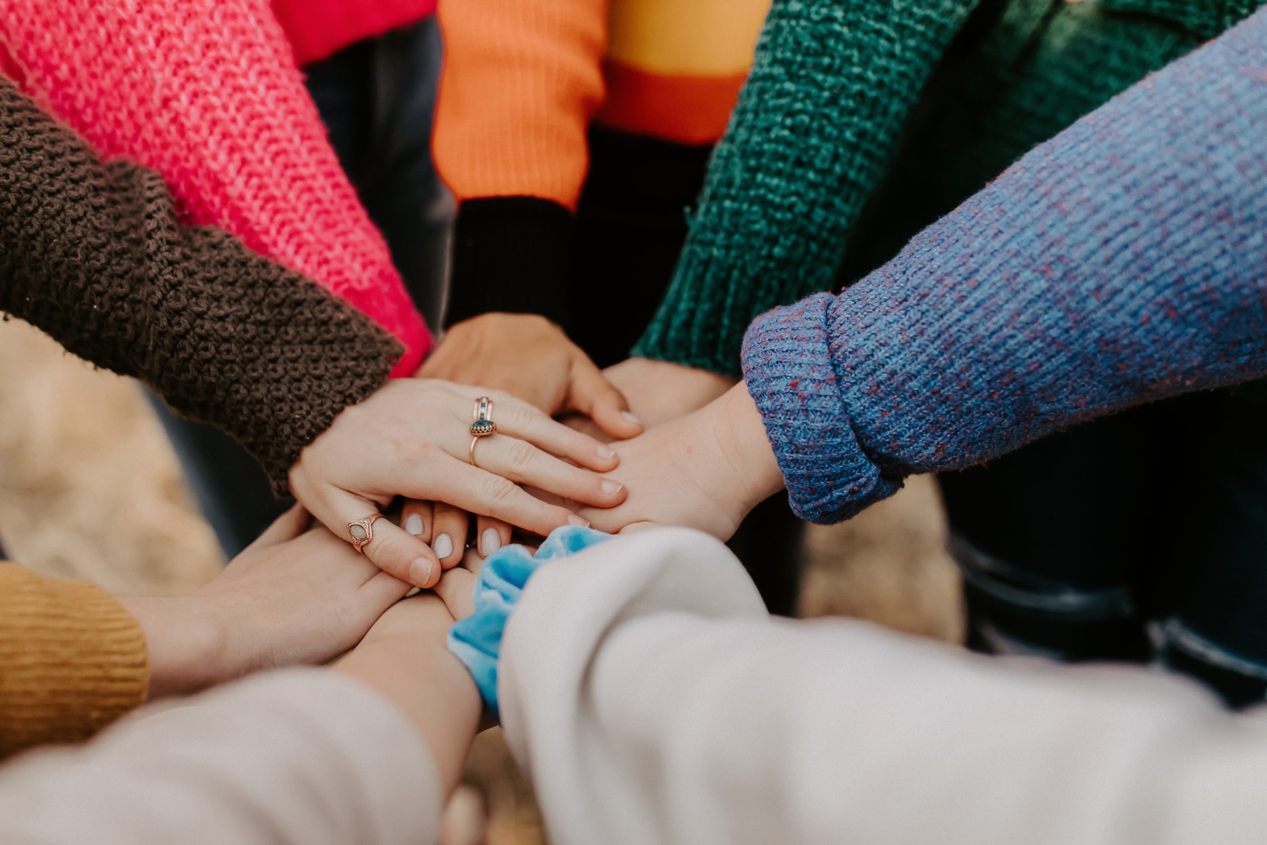 Stacked hands in a group circle, each person wearing a different colour sweater.