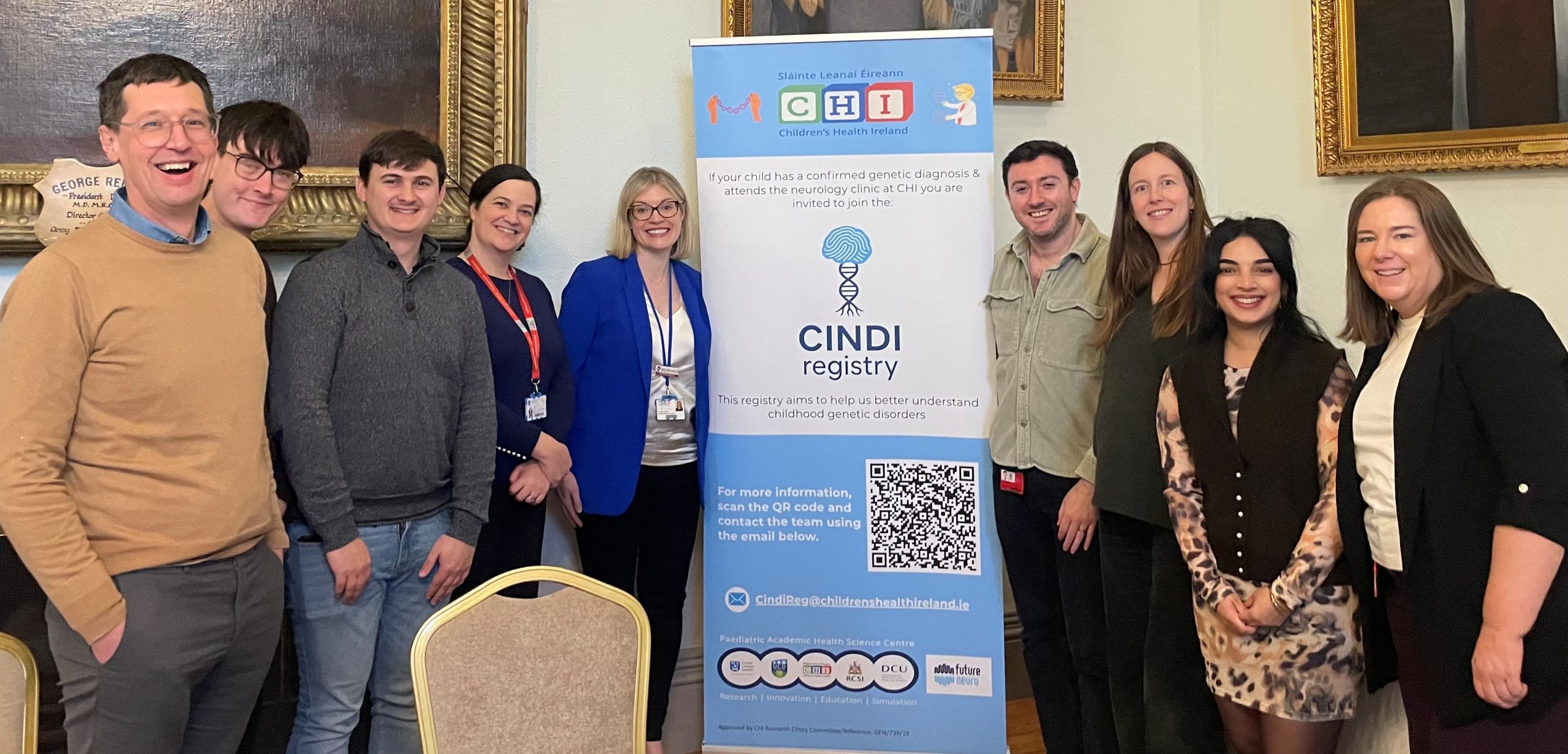 A group of people standing together in an indoor setting beside a tall CINDI Registry banner. The banner displays the CINDI Registry logo with a stylised DNA graphic, information about the registry, a QR code, and logos of partner organisations including Children’s Health Ireland and RCSI. The group is positioned in front of framed artwork on the wall, and chairs are visible in the foreground.