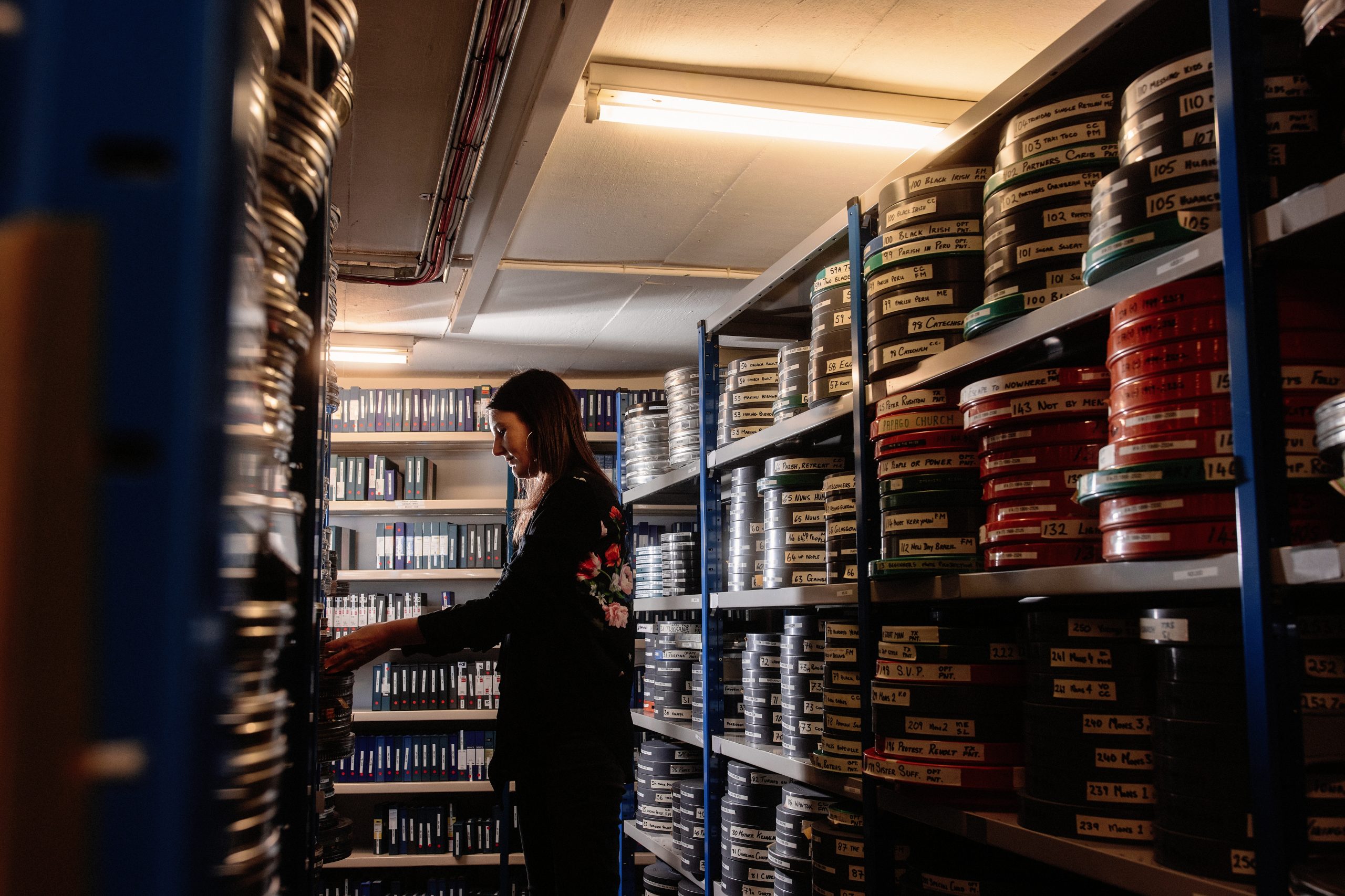 A woman stands between rows of metal shelving with labelled film reels, examining materials in a storage area with overhead lighting.