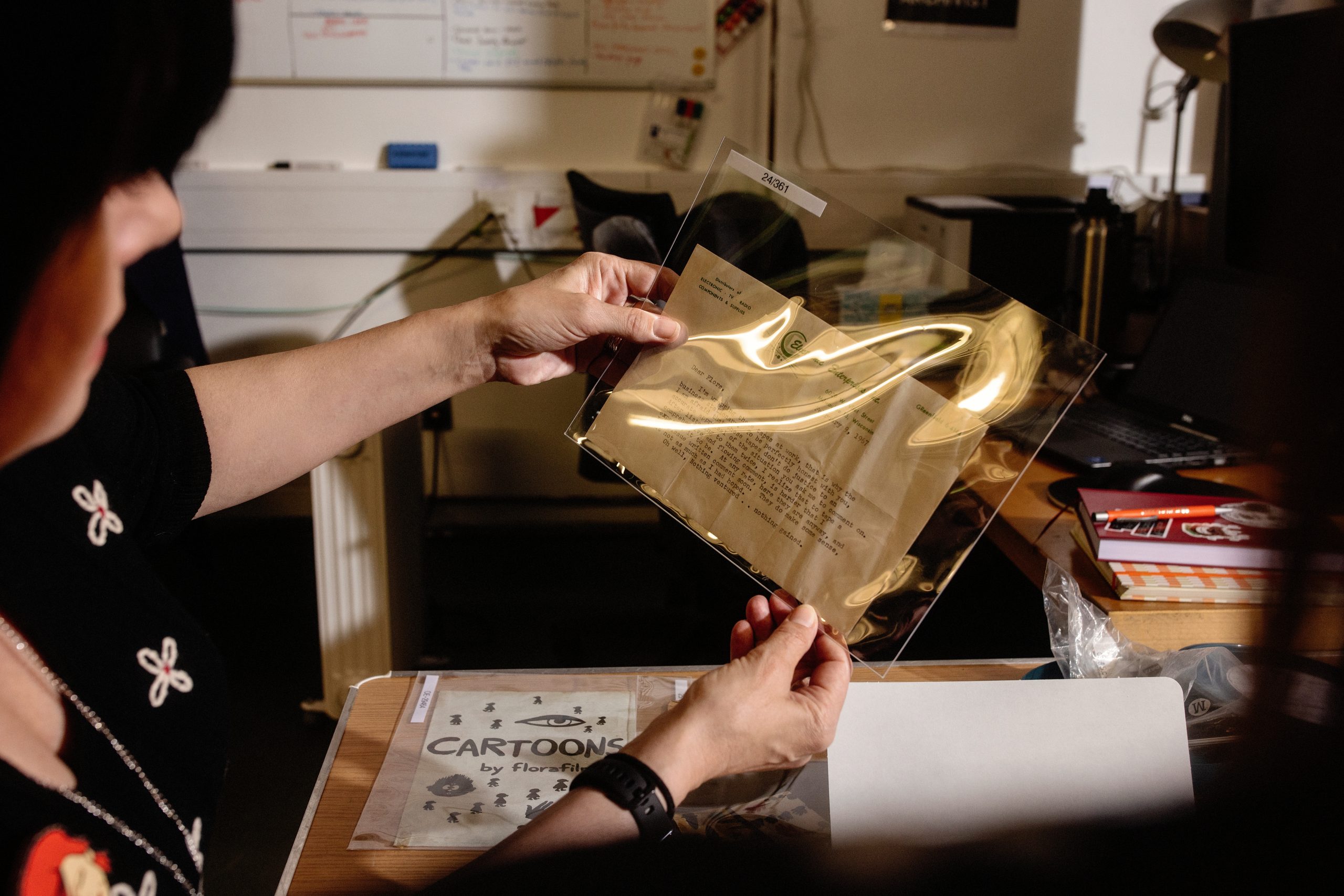 Hands holding a clear archival sleeve containing a typed document, with additional papers, a “Cartoons by Flora Kerrigan” folder, and archival materials spread out on the table beneath.