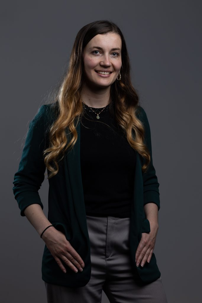 Studio portrait of a person with long wavy hair, wearing a dark top and green blazer, standing against a grey background.