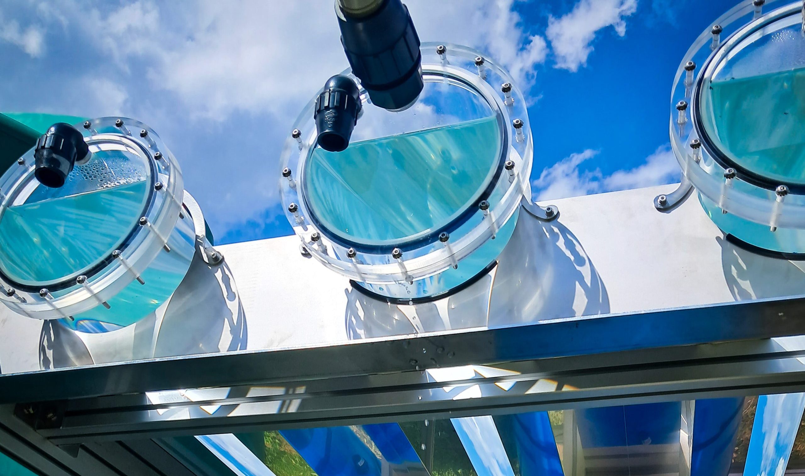Close-up view of an outdoor scientific or industrial setup with three transparent circular chambers partially filled with water, connected by black nozzles. The equipment’s reflective metal surfaces mirror the blue sky and clouds above