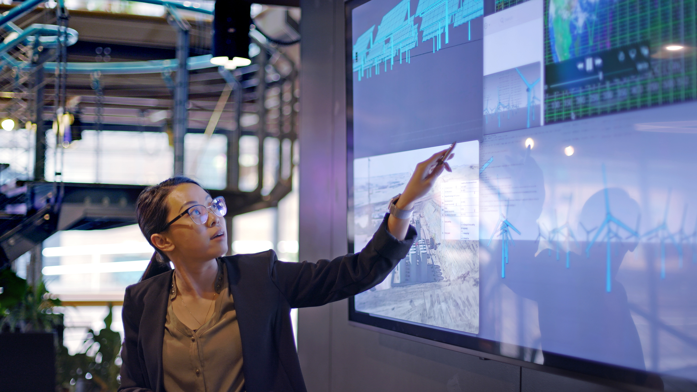 A photograph of a woman conducting a seminar with the aid of a large screen. The screen is displaying data concerning low carbon electricity production with solar panels and wind turbines.
