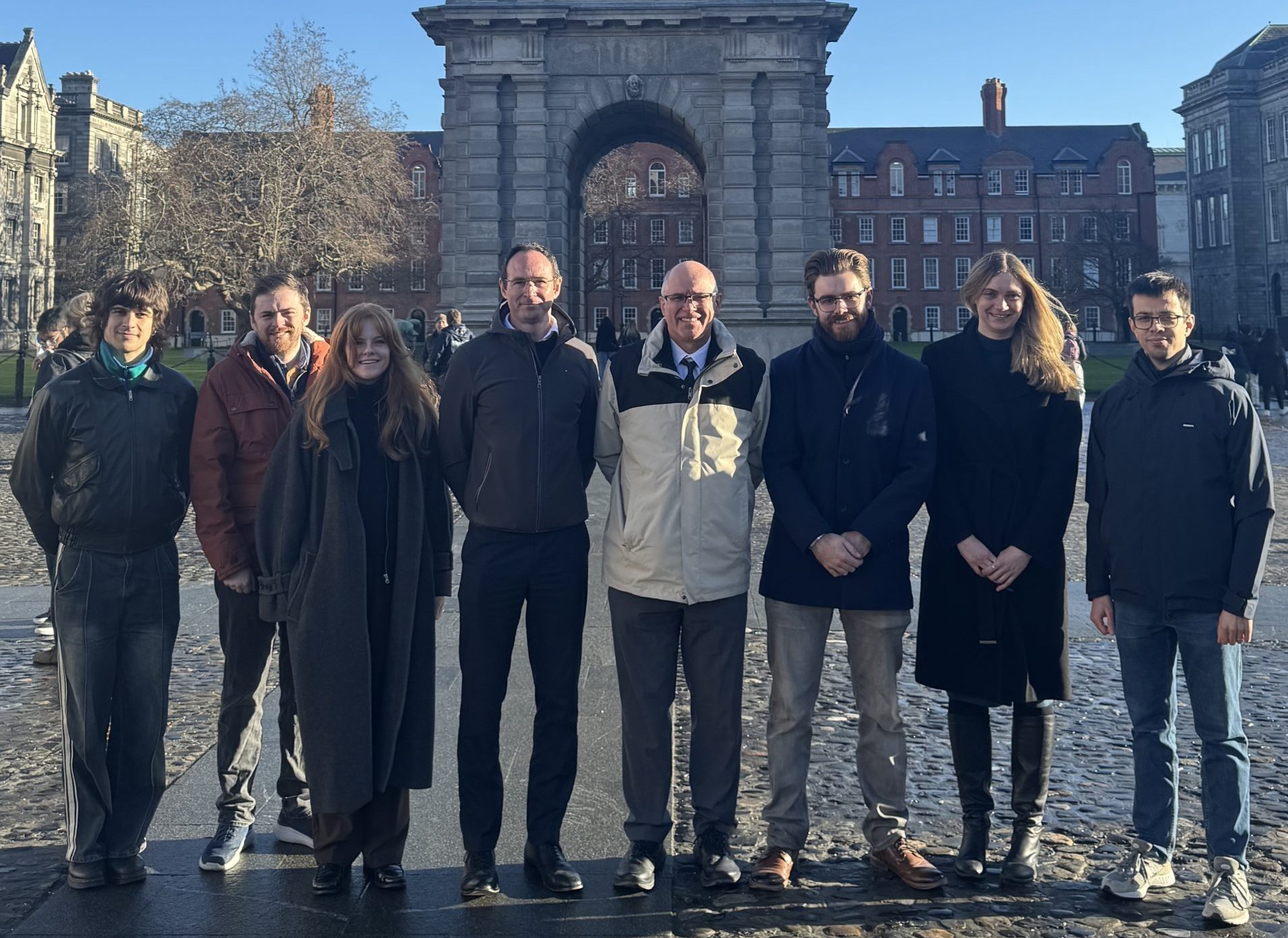 Eight people stand in a line in front of the Campanile at Trinity College Dublin, a historic bell tower with an arched base and domed top, set in a cobblestone courtyard under a clear blue sky.