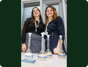 Photograph of two women standing behind a table displaying four glass volumetric flasks filled with granular material, each flask labelled with blue text indicating "200 ml."