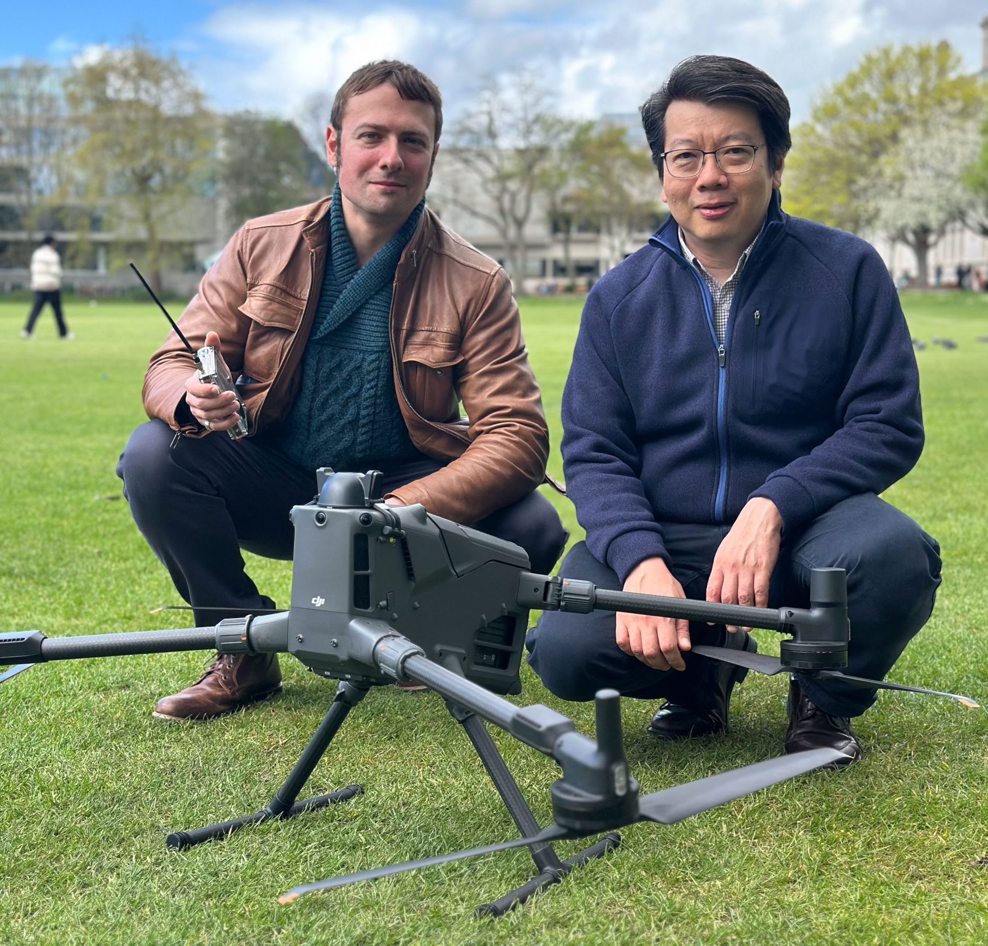 Two researchers kneel on a grass field beside a large drone examining its components. The scene shows a field test of drone technology.