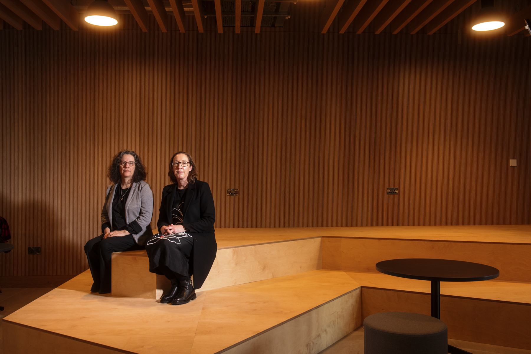 Two people sit on tiered wooden seating in a modern auditorium with a wood‑panelled backdrop. The space is set up for talks or presentations.