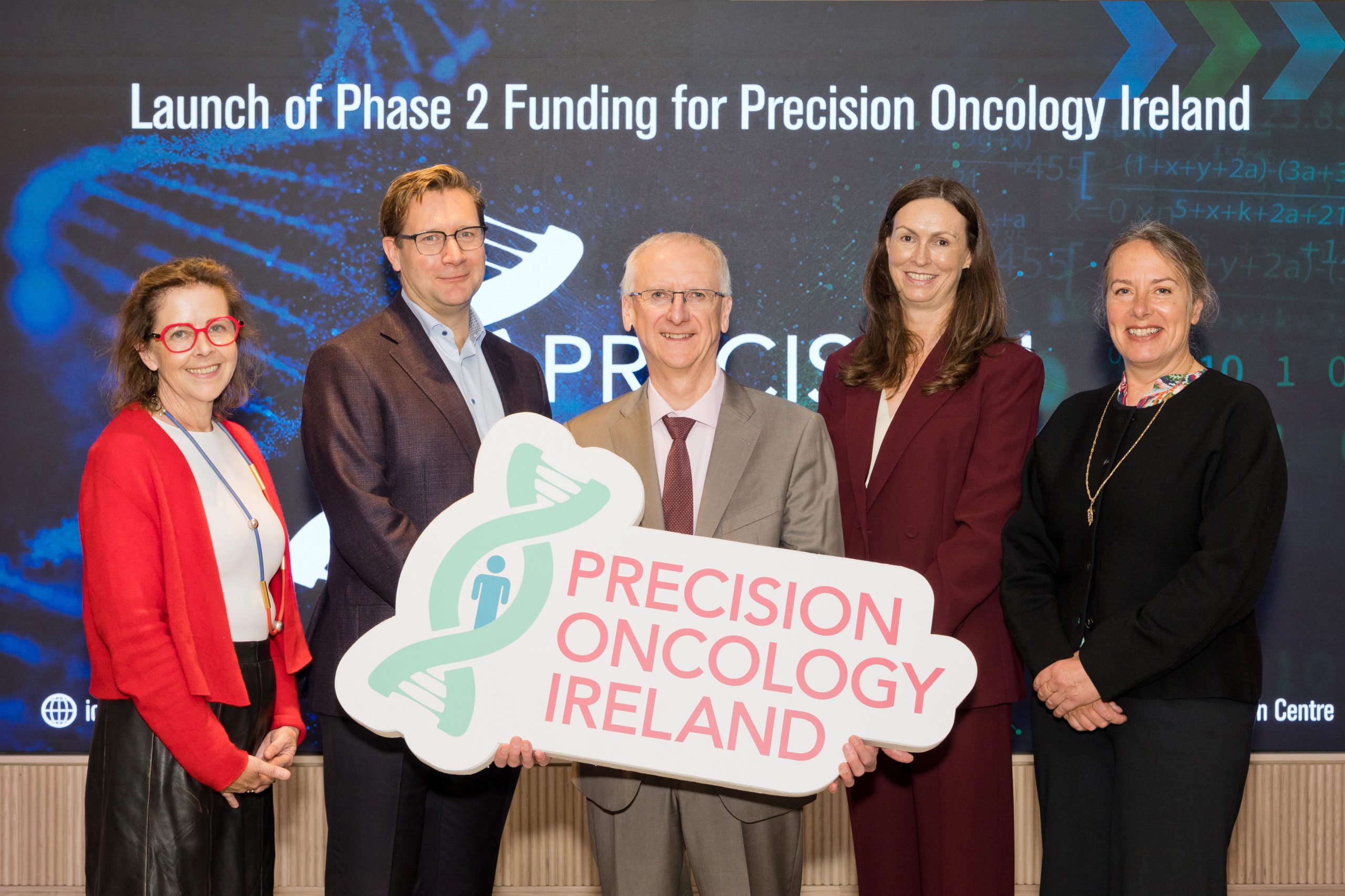 Five people stand on a stage holding a “Precision Oncology Ireland” sign in front of a presentation screen. The scene shows a funding announcement.