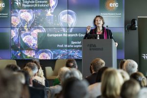 Maria Leptin stands at a podium addressing an audience during a conference forum, with a presentation screen displaying European Research Council branding behind.