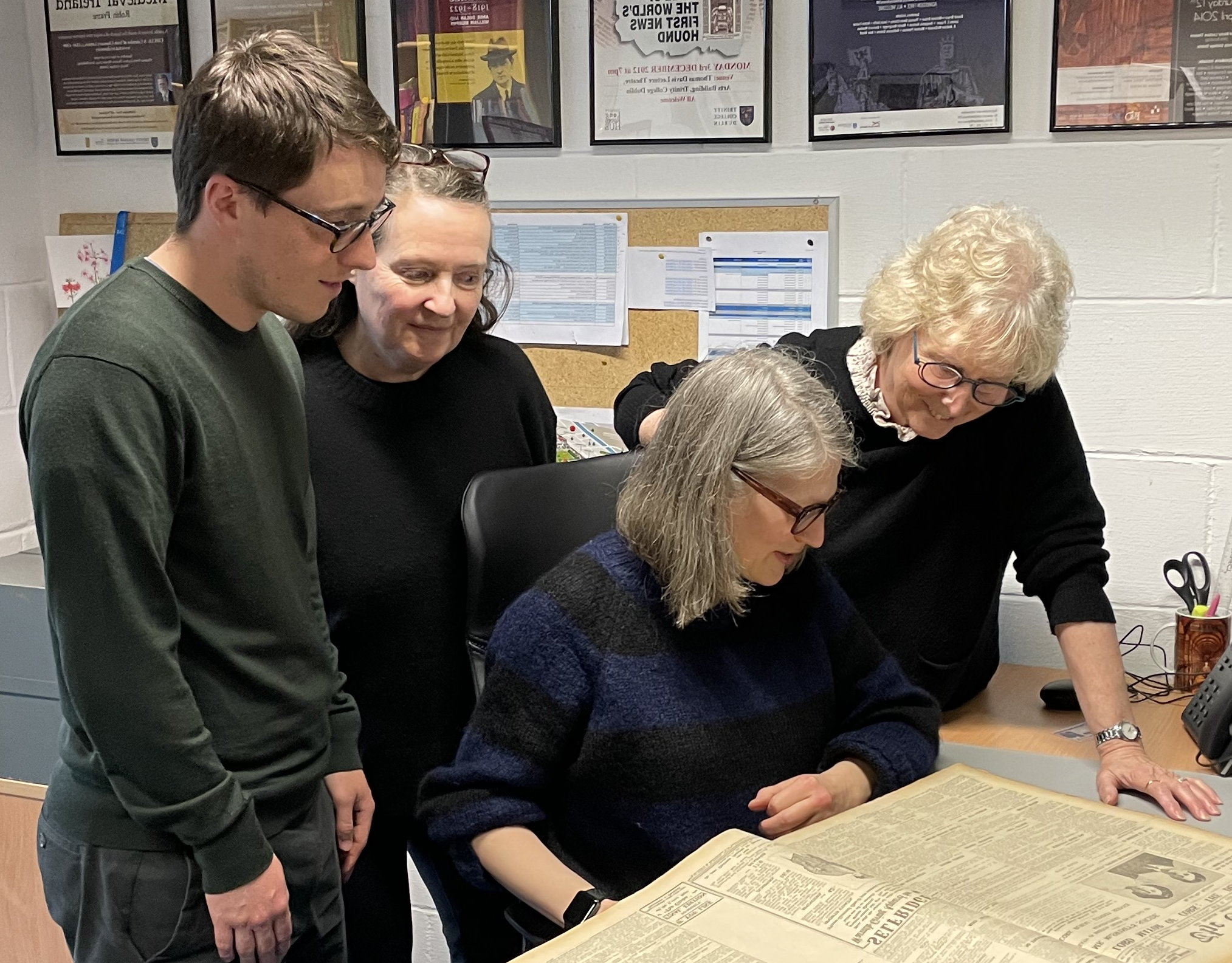 Four researchers examine a large historical document laid flat on a desk. The scene shows collaborative archival analysis.